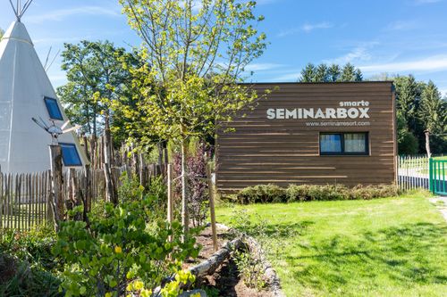 Holzgebäude mit Aufschrift "Smarte Seminarbox" in einem grünen Garten, daneben ein Tipi-Zelt und ein kleiner Zaun.