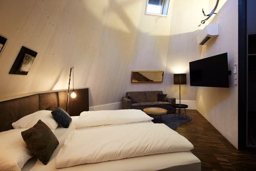 Doppelbett in modernem Schlafzimmer, flankiert von Nachttischlampen, Holzverkleidung, Sofa und Fernseher an der Wand.