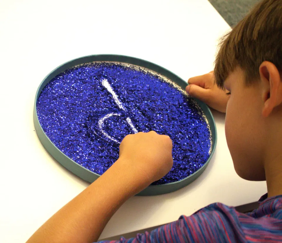 A pupil tracing a letter shape in a tray of blue glitter