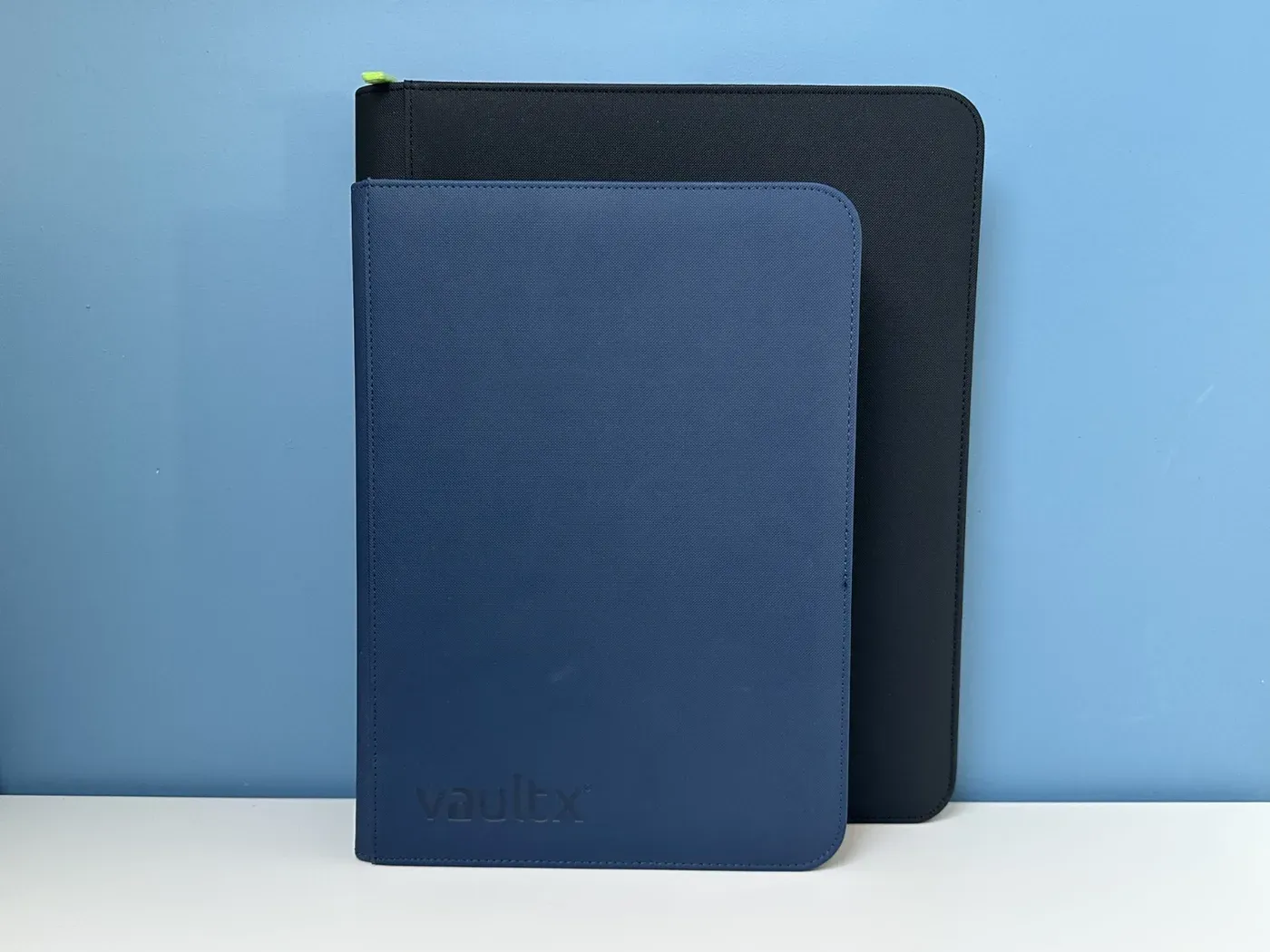 Twee 9-pocket binders van VaultX: vooraan een gewone blauwe binder met side-loading vakjes, erachter een zwarte toploader binder. De toploader binder is een stuk groter en neemt dus meer ruimte in.
