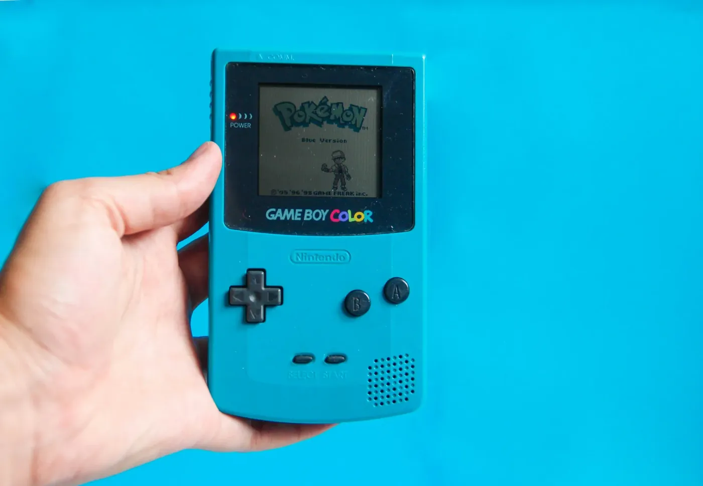 Hand met een turquoise Game Boy Color met het startscherm van Pokémon Blue op een blauwe achtergrond