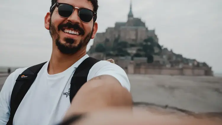 Man in sunglasses taking a selfie with a hilltop abbey behind him