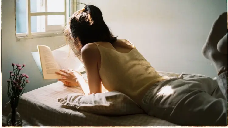 Woman lying on a bed reading a book in warm afternoon light