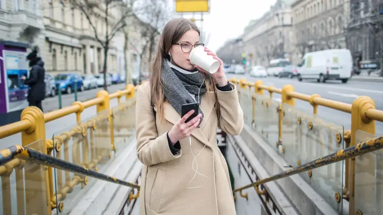 Young woman wearing glasses and earbuds sipping takeaway coffee.