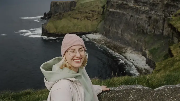 Smiling woman in a beanie hat standing above the Cliffs of Moher