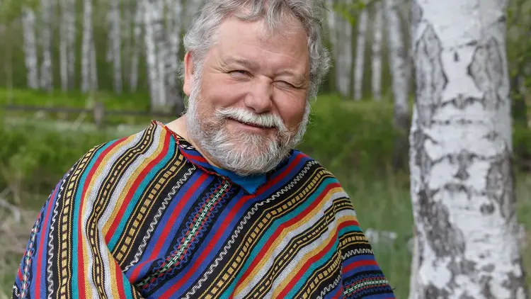 Older man in a colourful striped jumper smiling beside a birch tree