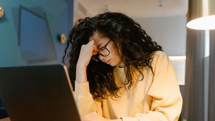 Woman in glasses rubbing her forehead while working at a laptop at night
