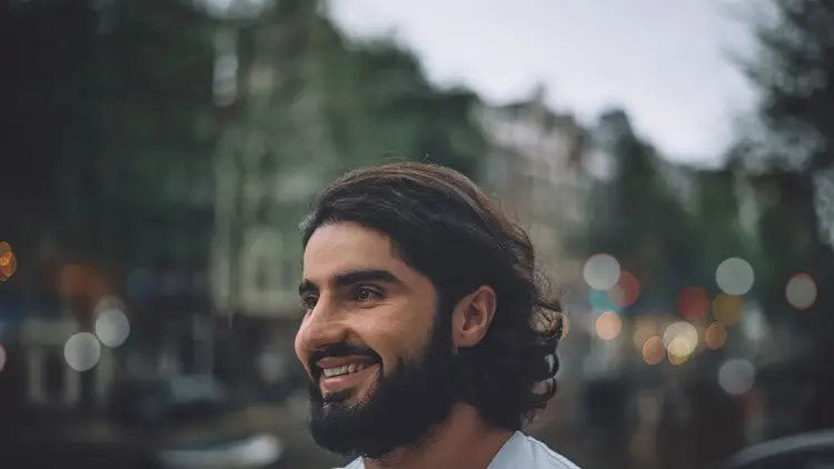 Bearded man smiling as he looks away, city lights blurred behind him