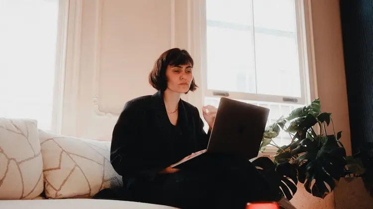 Woman sitting on a sofa by a bright window, working on a laptop