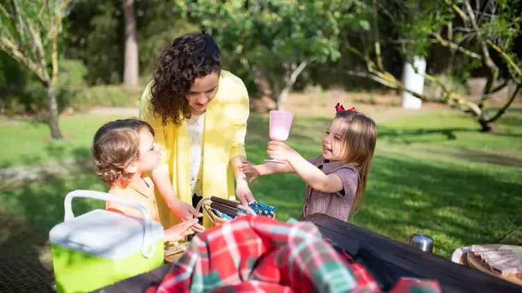 Woman handing a cup to a toddler at a picnic table in a park