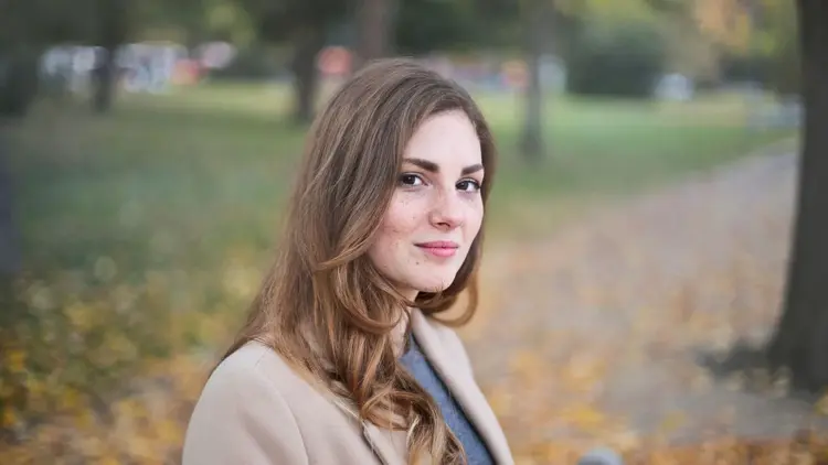 Woman in a camel coat standing in a park among fallen autumn leaves