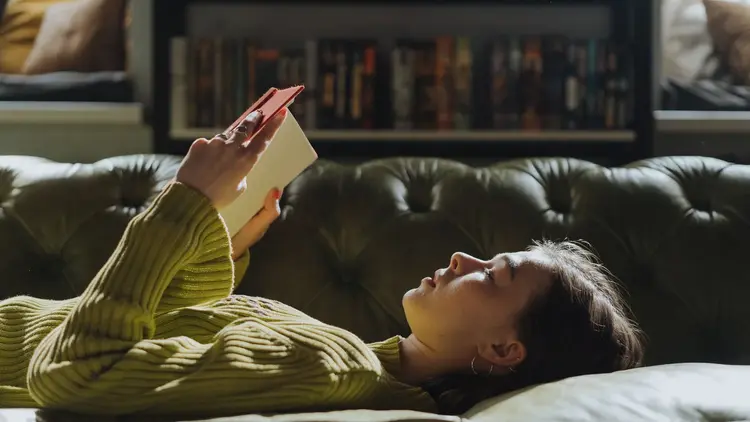 Woman lying back on a sofa reading, lit by a warm lamp
