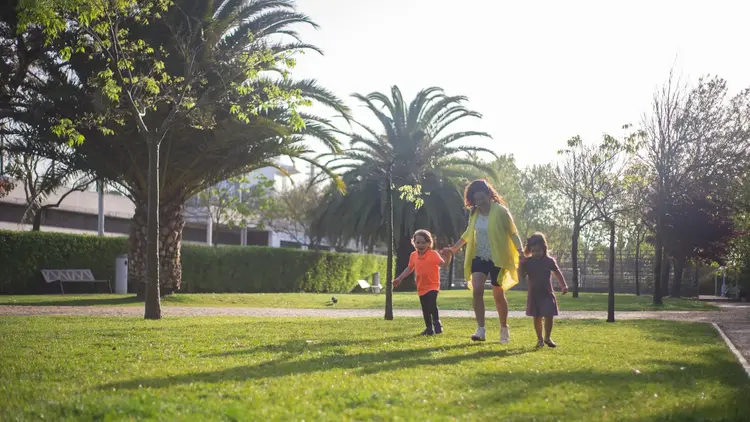 Woman walking across a sunlit park lawn with two young children