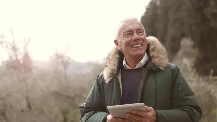 Older man laughing while holding a tablet on a country walk