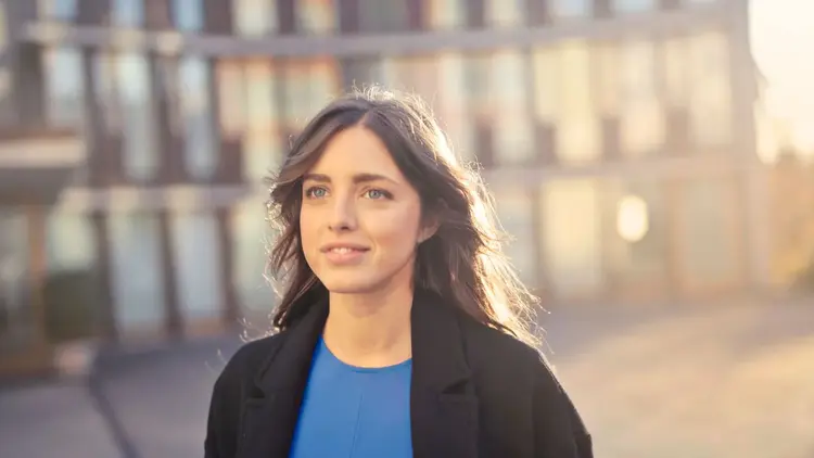 Woman in a dark coat smiling as she walks along a sunlit city street