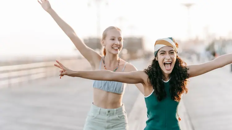 Two young women laughing with their arms outstretched on a promenade