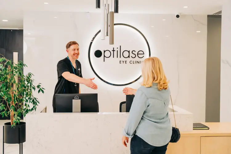 A smiling staff greets a female patient at the modern white front desk of Optilase Eye Clinic.