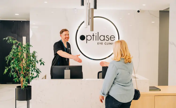 A smiling receptionist greets a female patient at the modern white marble front desk of Optilase Eye Clinic.
