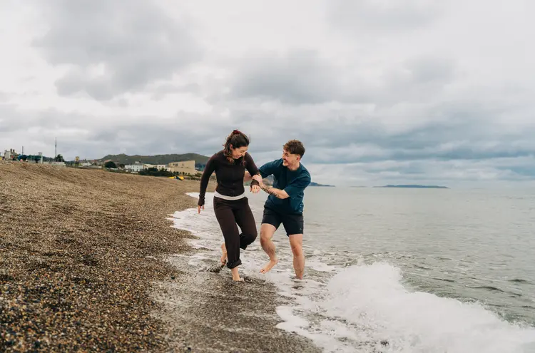 Two people laughing as they run barefoot along a rocky beach