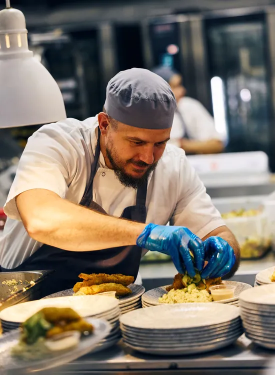 A chef wearing a gray cap and blue gloves carefully plating a dish in a professional kitchen with stacks of plates nearby.