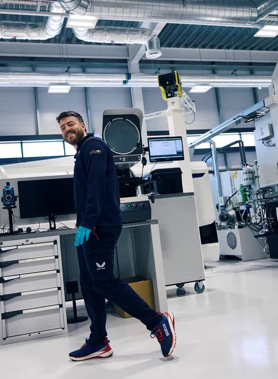 A man in athletic gear and gloves smiles while walking inside a modern industrial facility with machinery and equipment.