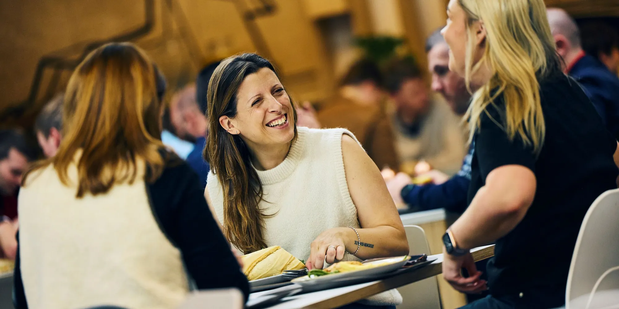 Three people sitting at a table, smiling and talking while enjoying a meal in a lively indoor setting.
