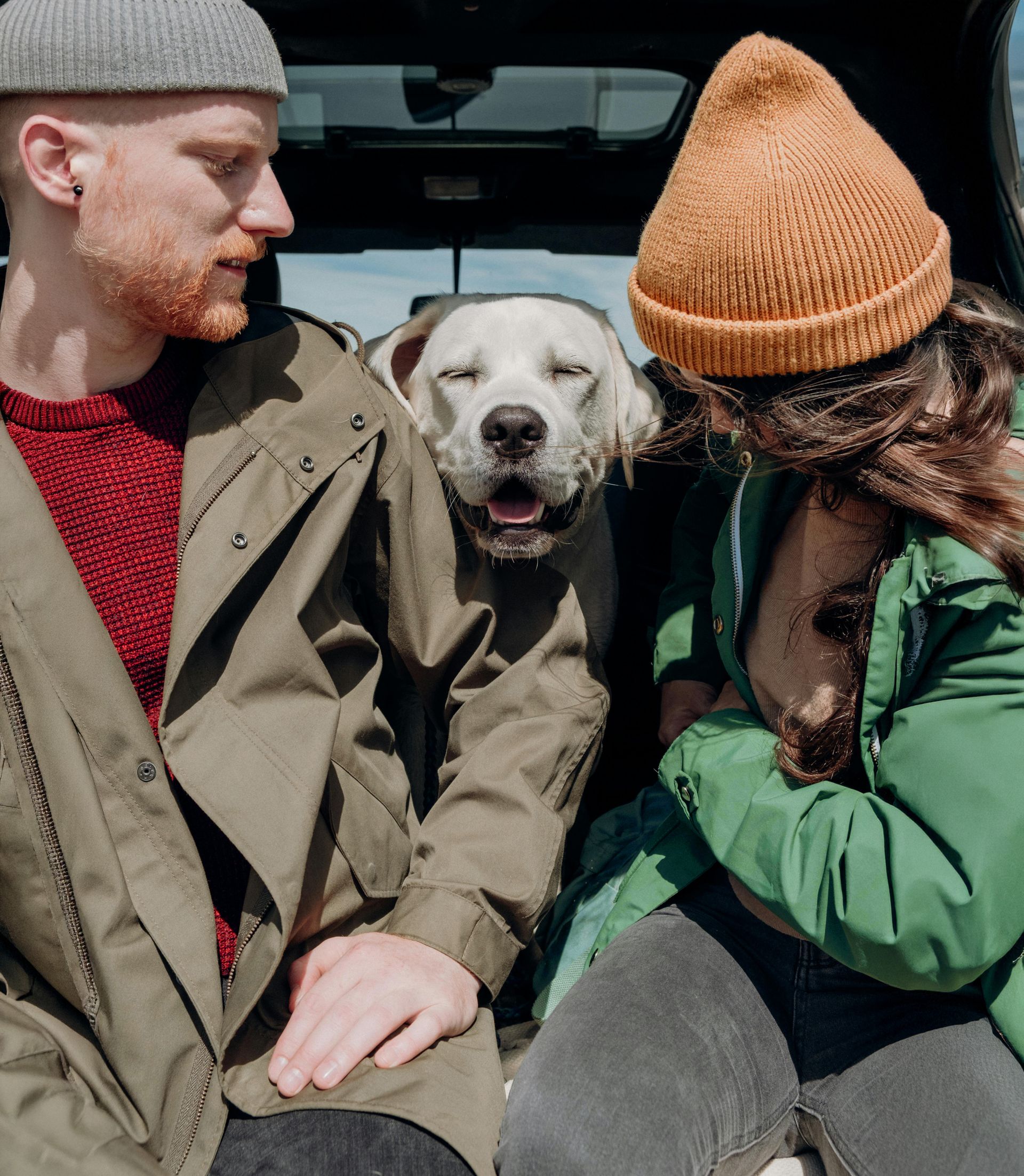 Ein Mann und eine Frau mit Mützen sitzen in einem Fahrzeug, zwischen ihnen ein fröhlicher Hund, und genießen einen sonnigen Ausflug.