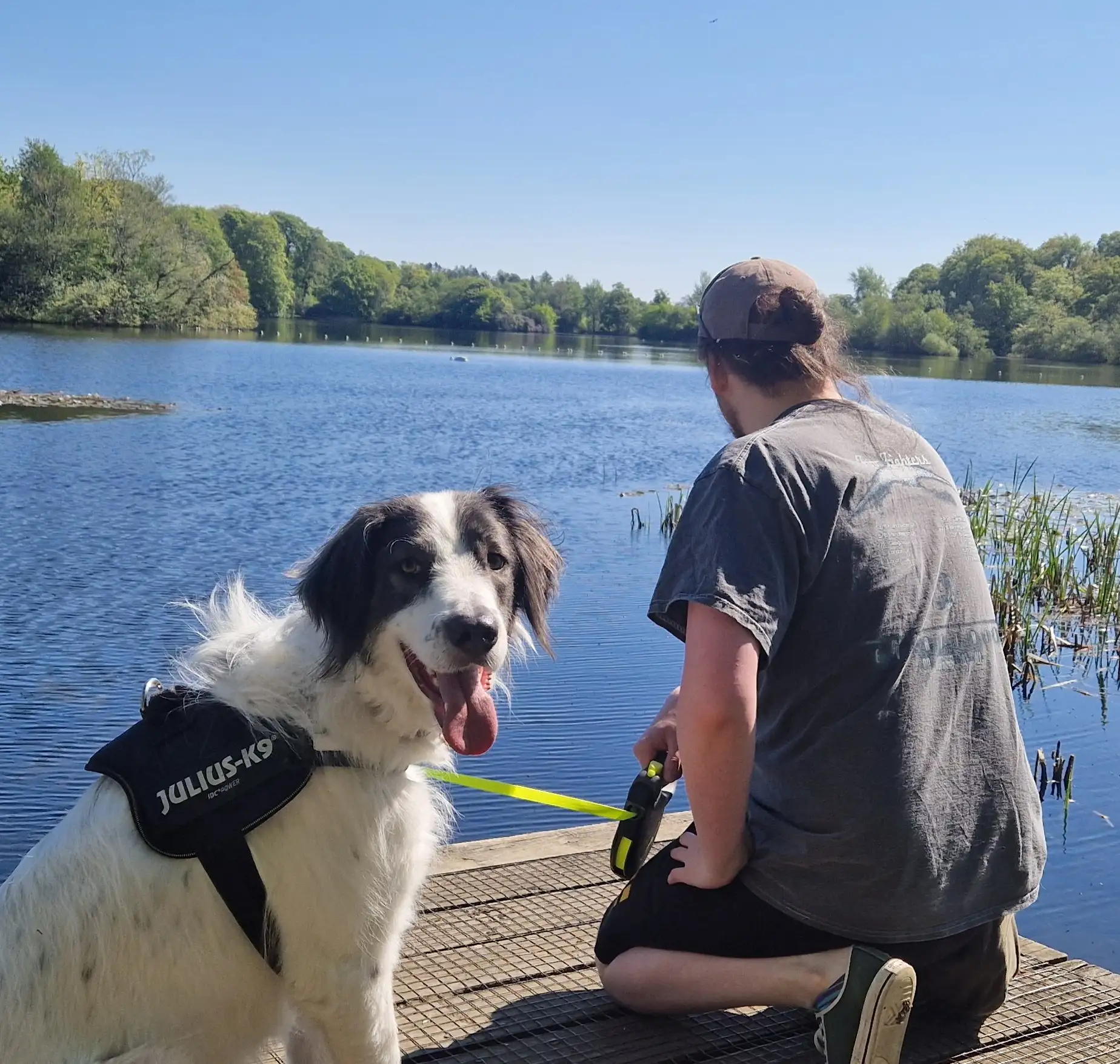 A picture of Ollie Smith kneeling and looking out onto a lake with his dog, Moose, a large black-and-white dog looking happily at the camera with an open mouth and his tongue hanging out.