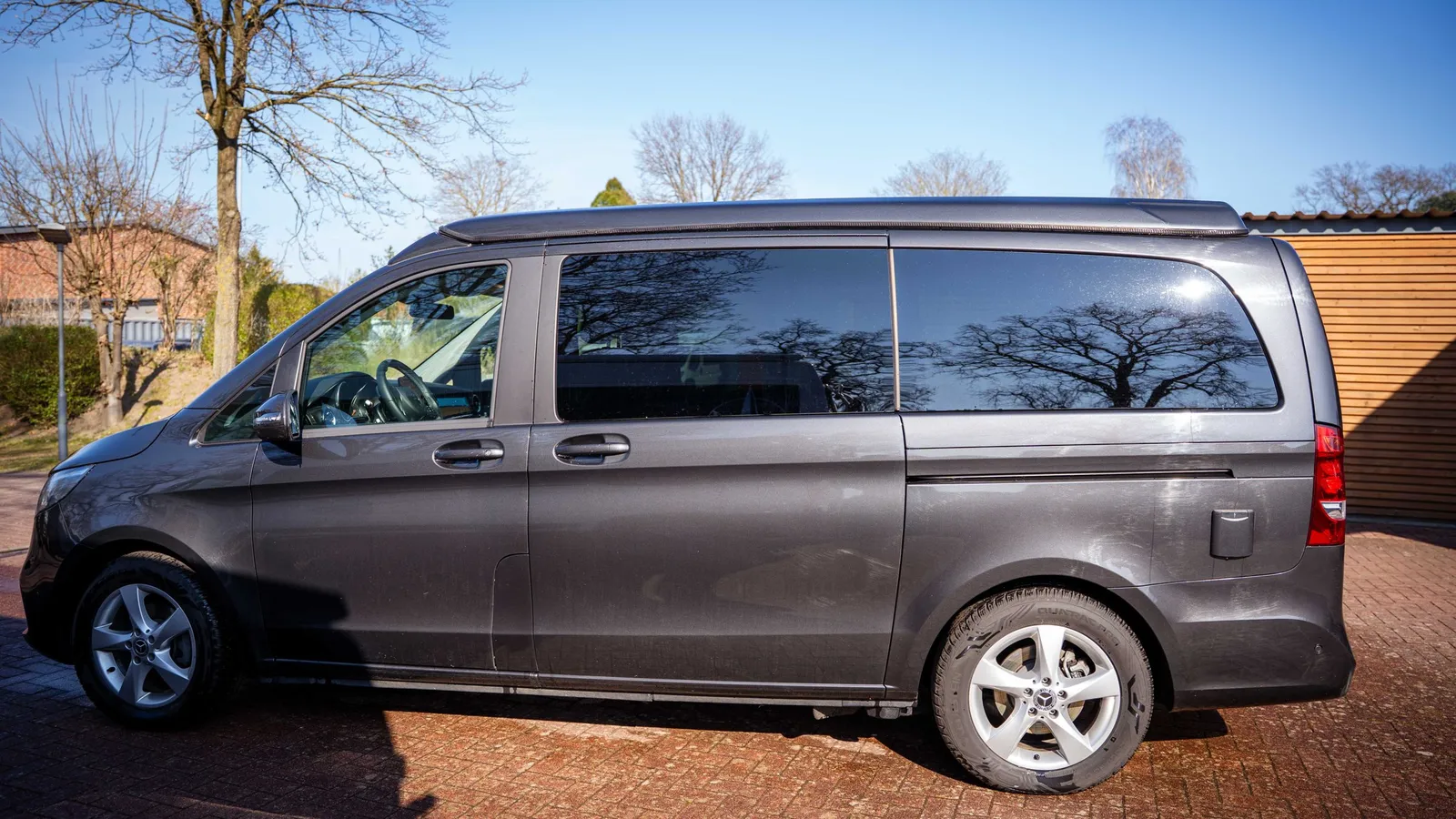 Vanstar plus – Grauer Pössl Vanline Vanstar plus Campervan in Seitenansicht auf einer gepflasterten Auffahrt unter blauem Himmel.