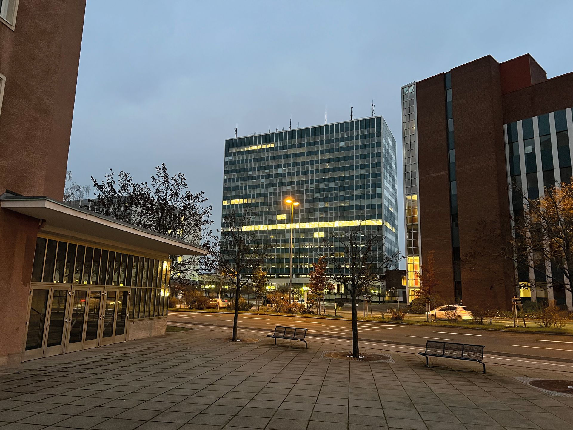 Unbearbeitete Aufnahme desselben Bürohochhauses in der Dämmerung mit grauem Himmel und nur vereinzelt beleuchteten Fenstern