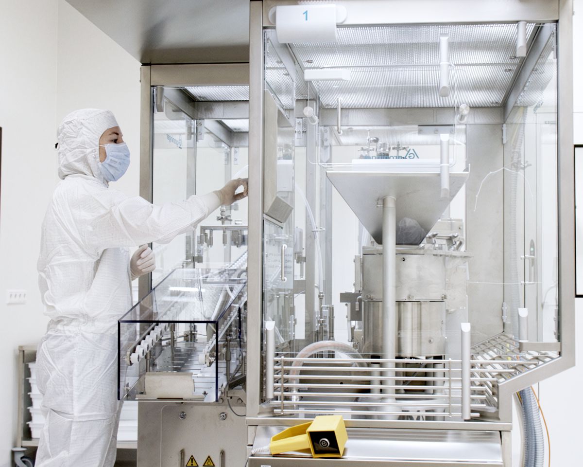 Mitarbeiterin in weißem Reinraumanzug mit Mundschutz bedient eine Abfüllanlage hinter Glas
