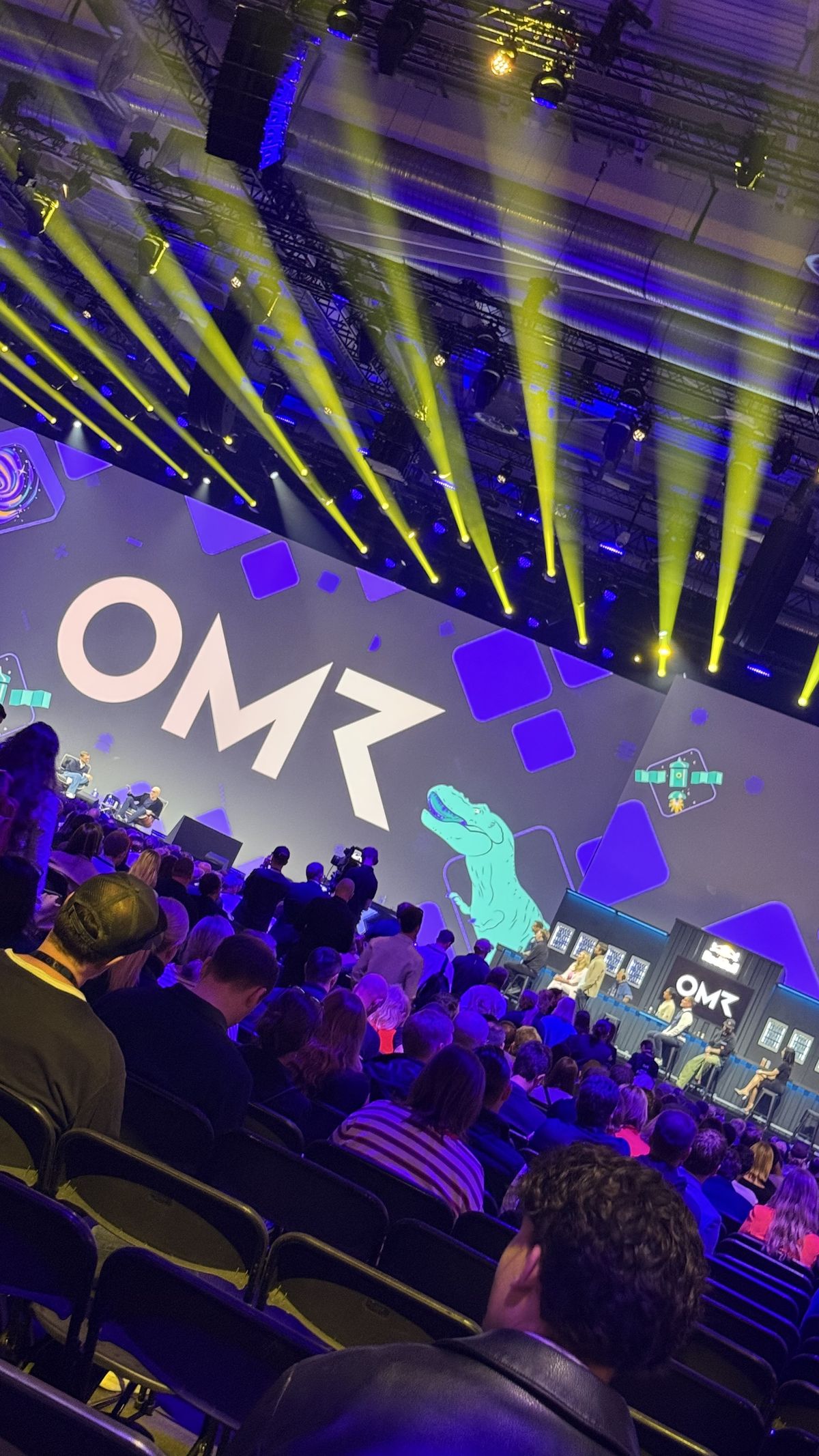 Blick aus dem Publikum auf die OMR Stage mit großem OMR-Schriftzug, Dinosaurier-Grafik und gelben Scheinwerferstrahlen, schräge Aufnahme
