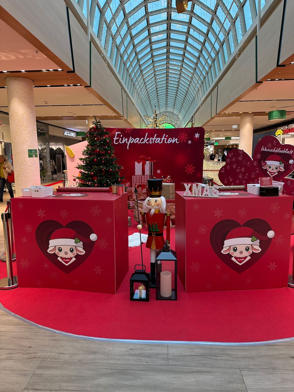 Weihnachtliche Einpackstation im FLAiR Fürth unter dem Glasdach: rote Theken mit FLAiRY im Herz, Tannenbaum, Nussknacker-Figur und Wunschkleeblatt-Wand