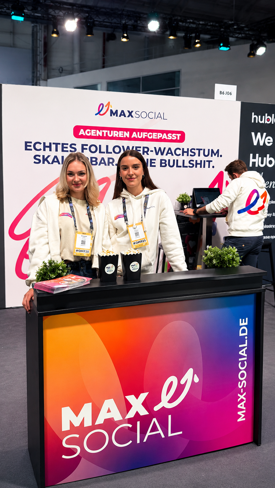 Zwei Mitarbeiterinnen in weißen MAXSOCIAL Hoodies hinter der Theke im Farbverlauf am Messestand B6 J06, dahinter die Rückwand mit Claim