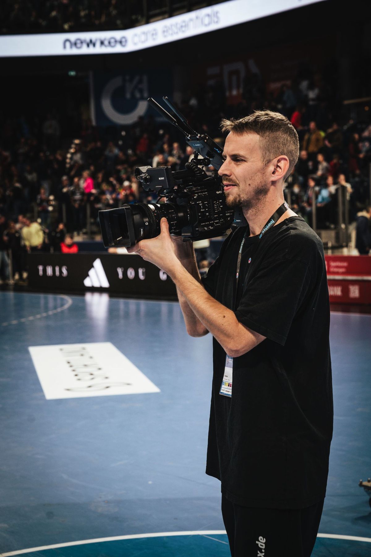Kameramann von fourplex filmt mit einer Sony-Kamera am Spielfeldrand der Handballarena während eines Heimspiels vor vollen Rängen