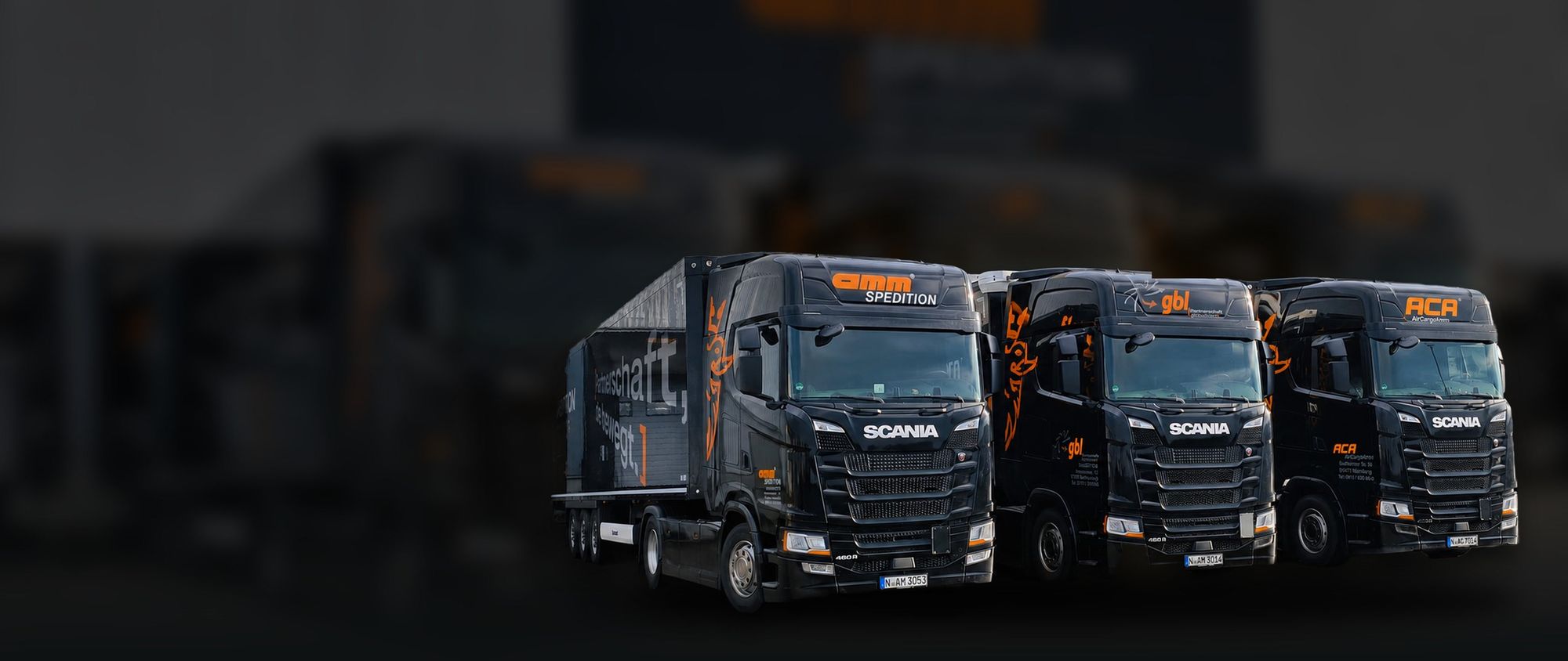 Drei schwarze Scania-Sattelzüge der amm Spedition mit orangefarbener Beschriftung vor dunklem Hintergrund