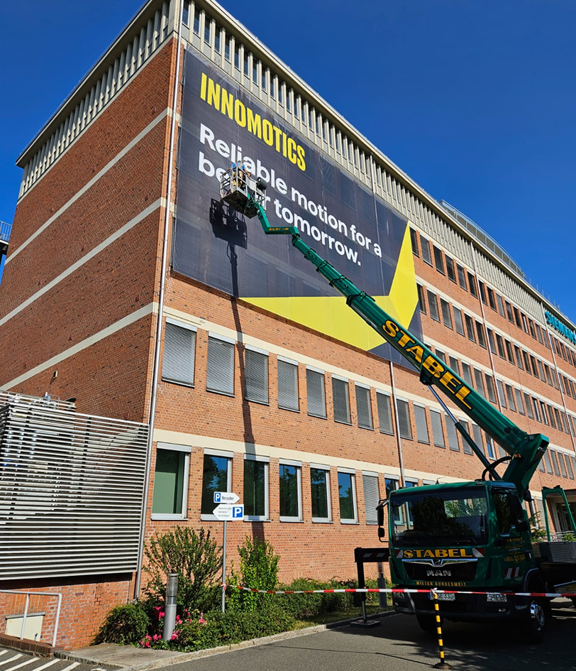 Hubsteiger montiert ein riesiges Innomotics-Banner „Reliable motion for a better tomorrow.“ an der Backsteinfassade eines Bürogebäudes vor blauem Himmel