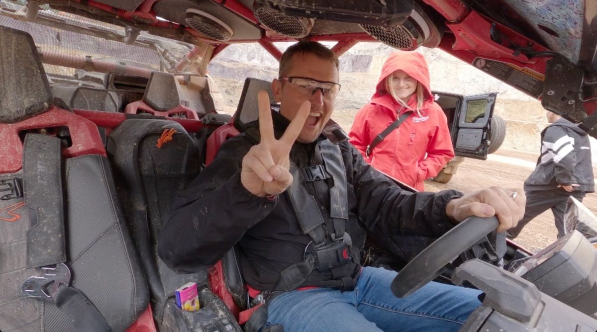 Lachender Fahrer mit Schutzbrille zeigt im Offroad-Buggy das Peace-Zeichen, im Hintergrund eine Frau in roter Milwaukee-Jacke