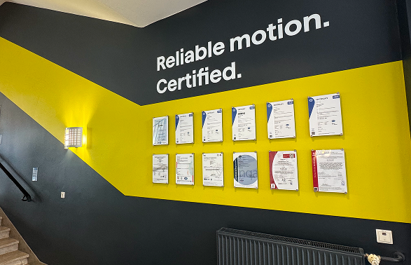 Treppenhaus in Anthrazit und Gelb mit Schriftzug „Reliable motion. Certified.“ und zwölf gerahmten Zertifikaten an der Wand