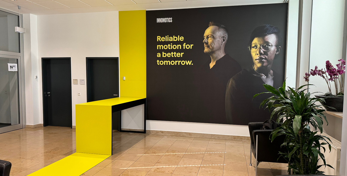 Foyer mit großem Wandbild zweier Personen und dem Claim „Reliable motion for a better tomorrow.“, davor ein gelber Stehtisch mit gelbem Bodenstreifen