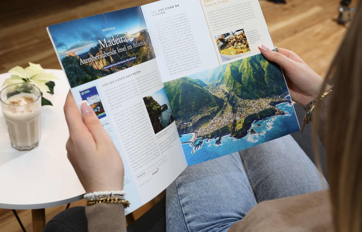 Frau hält das aufgeschlagene Reiselust-Magazin mit Doppelseite über Madeira und Küstenpanorama, daneben ein Glas Latte macchiato