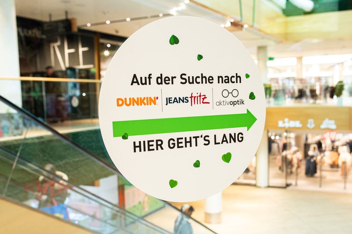 Runde Wegeleit-Beklebung an der Glasbrüstung im FLAiR Fürth: „Auf der Suche nach Dunkin', Jeans Fritz, aktivoptik – hier geht's lang“ mit grünem Pfeil, dahinter die Rolltreppe