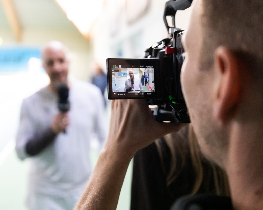 Kameramann filmt einen Coach mit Mikrofon in einer Tennishalle, Blick über die Schulter auf das Kameradisplay