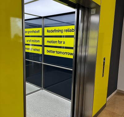 Geöffneter Aufzug mit gelber Verkleidung und gelben Folienstreifen „Redefining reliable motion for a better tomorrow“ an der verspiegelten Kabinenwand