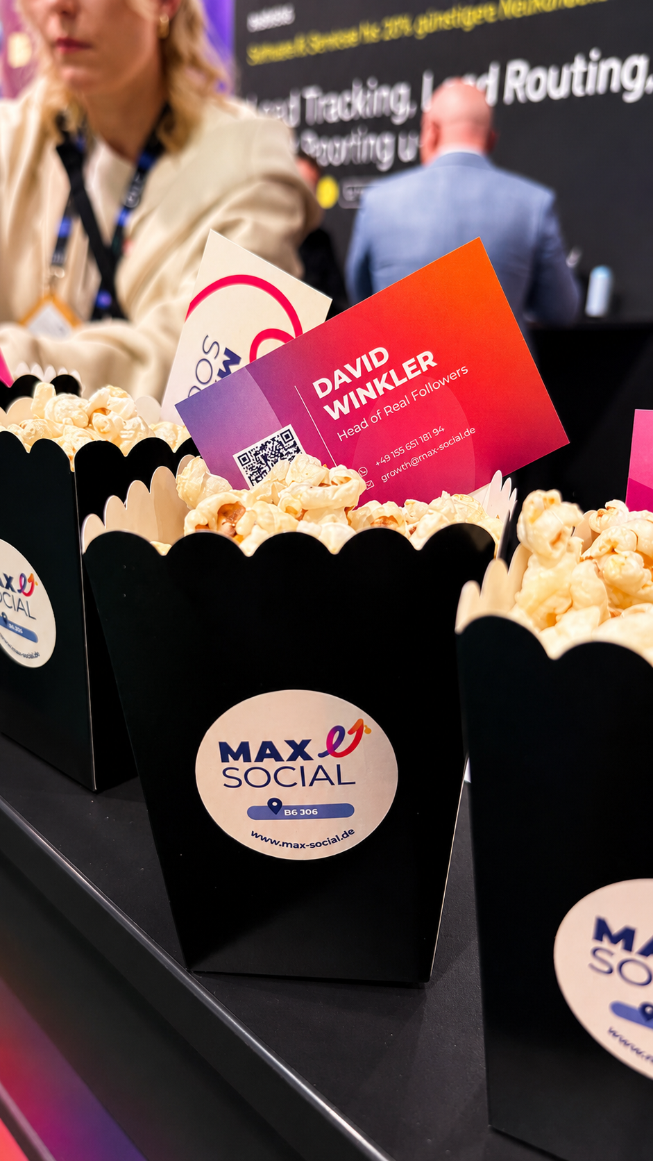 Schwarze Popcorntüten mit MAXSOCIAL Sticker und Standnummer B6 J06, dazwischen eine Visitenkarte im Farbverlauf, im Hintergrund Messebetrieb