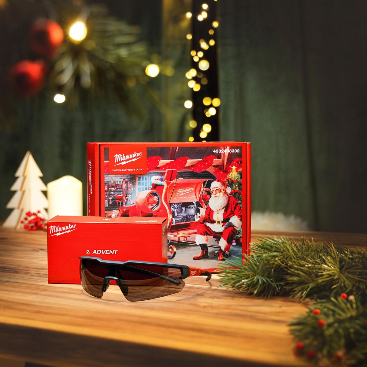 Milwaukee-Adventskalender mit Weihnachtsmann-Motiv auf Holztisch, davor die Box „2. Advent“ und eine getönte Schutzbrille, dekoriert mit Tannenzweigen und Kerze