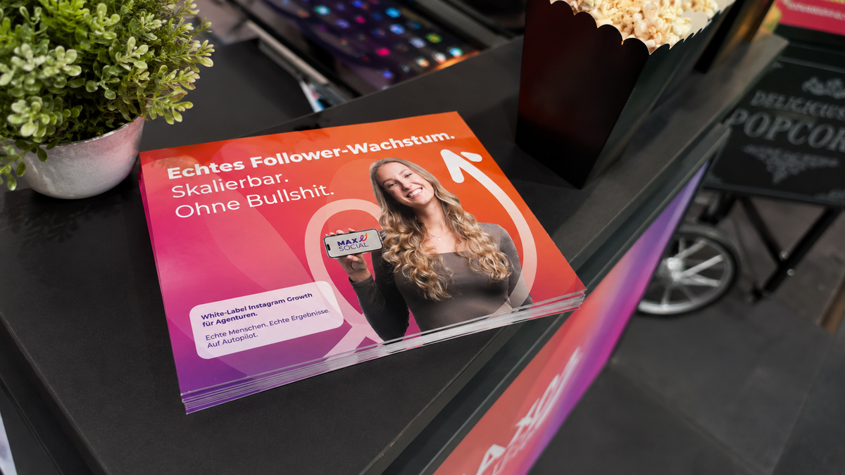 Mockup der MAXSOCIAL Broschüre im A5-Querformat mit Claim „Echtes Follower-Wachstum.“ und lächelnder Frau mit Smartphone, auf dunkler Messetheke neben Popcorn