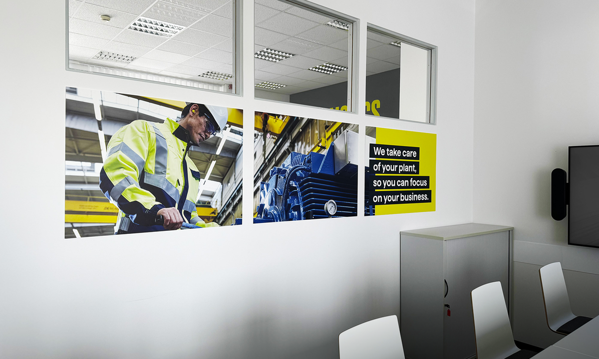 Besprechungsraum mit folierten Glasscheiben: Arbeiter mit Schutzhelm und Warnjacke, blaue Industriemaschine und Schriftzug „We take care of your plant, so you can focus on your business.“