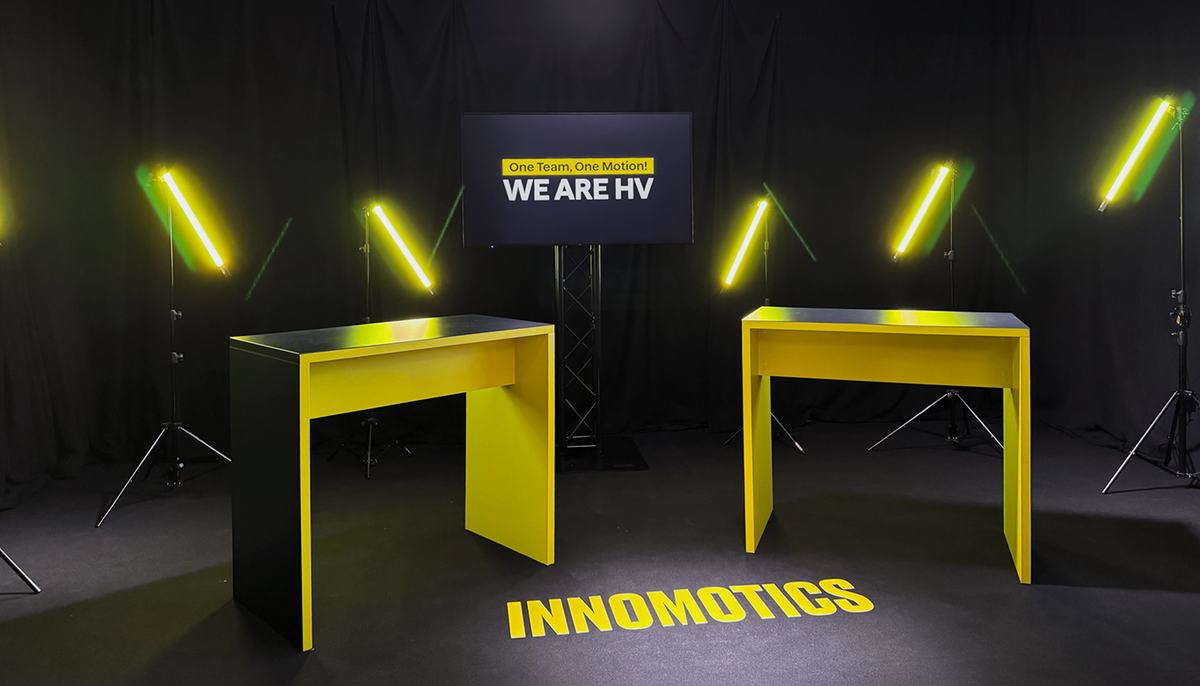 Studio-Set für das Townhall-Meeting: zwei gelbe Stehtische, Monitor mit „One Team, One Motion! WE ARE HV“, gelbe Leuchtstäbe und Innomotics-Schriftzug auf dem Boden