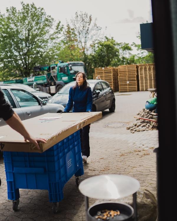 Frau schiebt eine blaue Rollbox mit großem Karton über den gepflasterten Firmenhof, im Hintergrund Paletten und ein Lkw